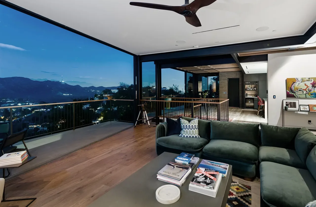 Living room with floor-to-ceiling glass opening to an exterior deck with railing and mountain view at dusk.