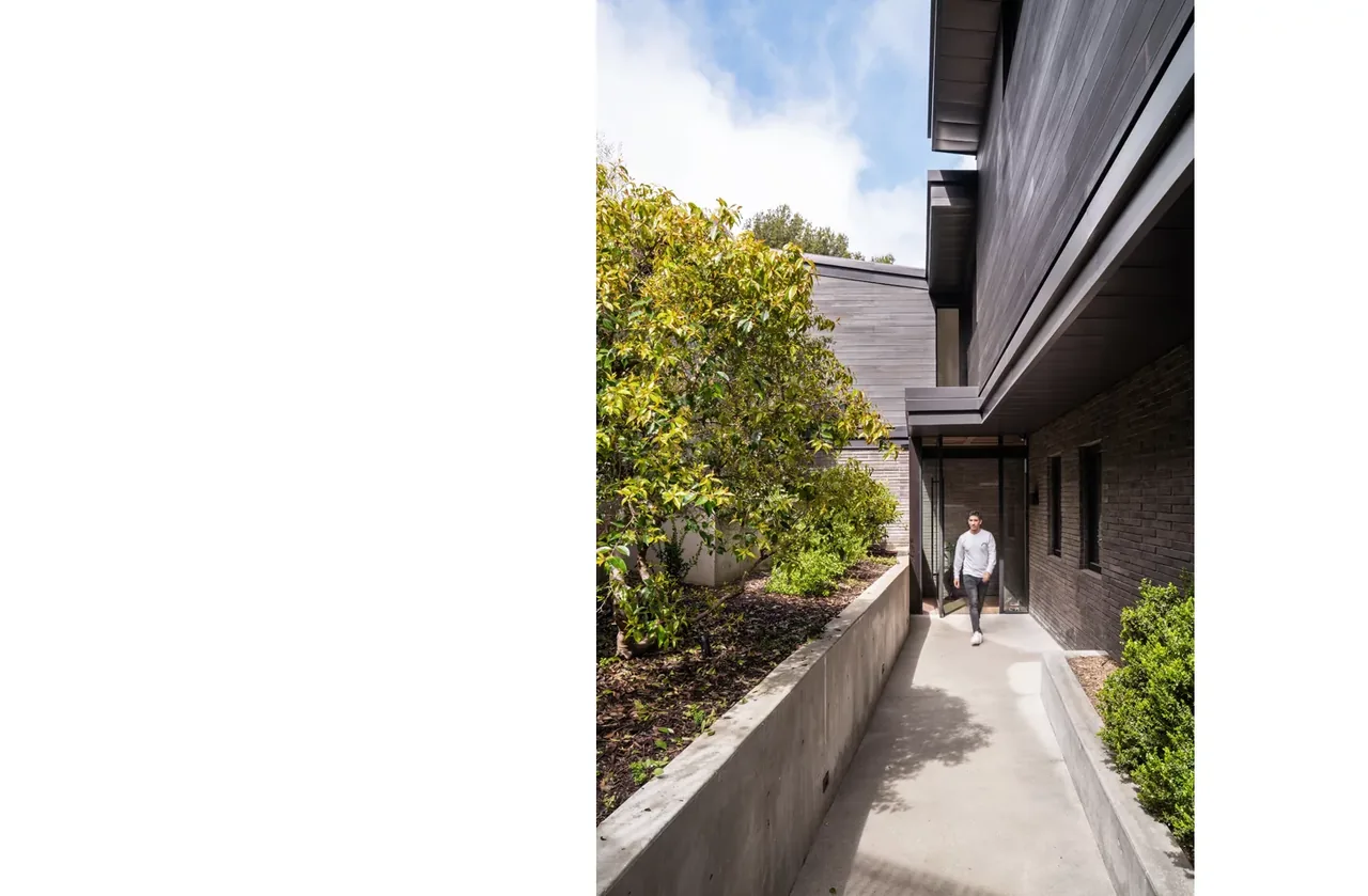 Narrow exterior pathway beside a modern house with concrete planters and wood siding.