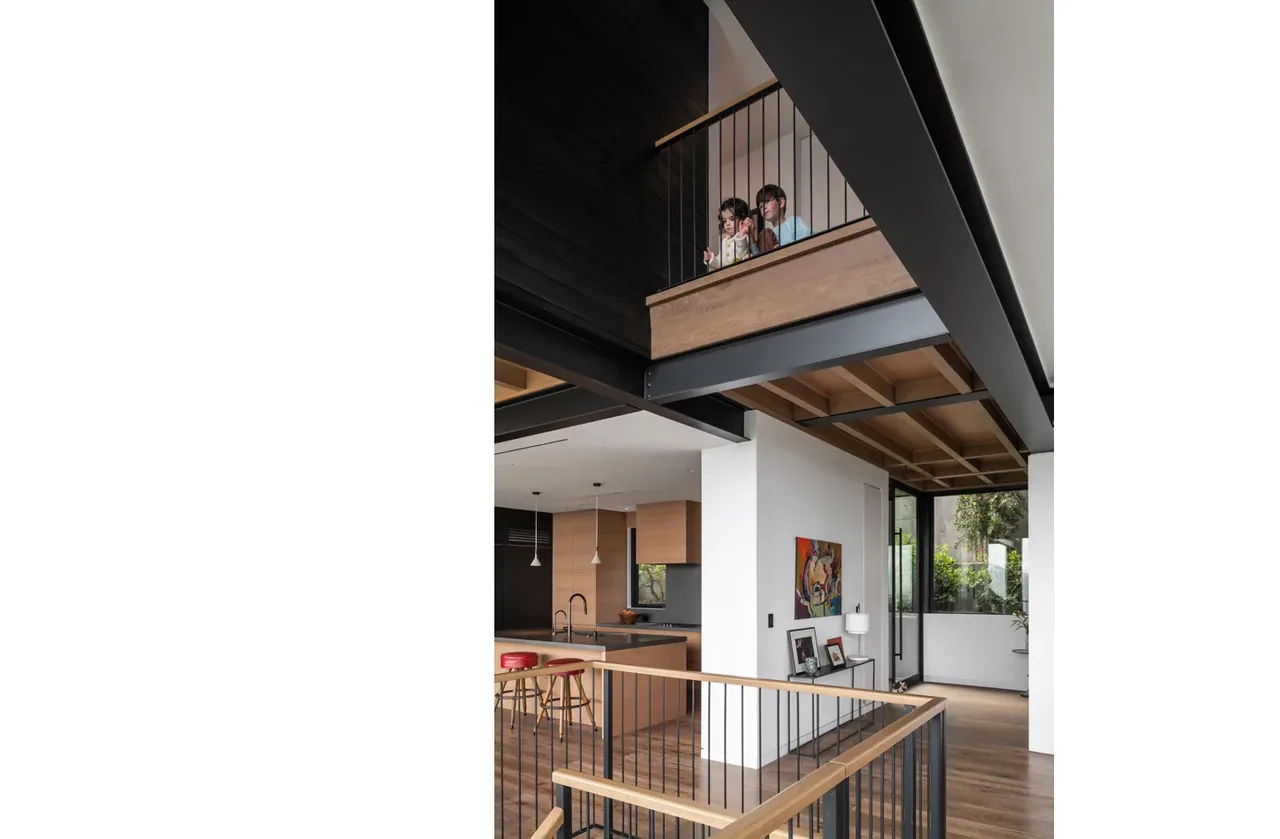 Two-level interior space with wood flooring, exposed wood ceiling beams, and a kitchen area under the upper floor balcony with metal railings.