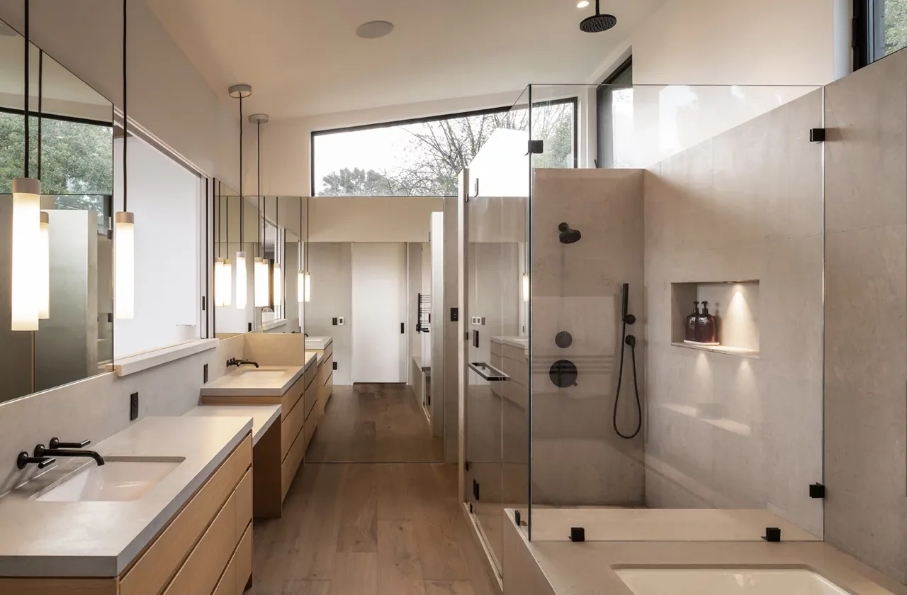 Bathroom featuring dual vanities with wooden cabinets, a glass shower enclosure, and a rain shower head beneath a high window.