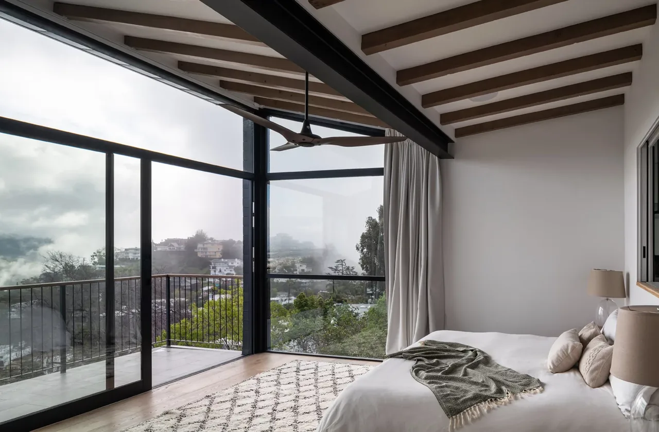Corner bedroom with exposed wood beam ceiling and floor-to-ceiling glass corner windows opening to balcony.