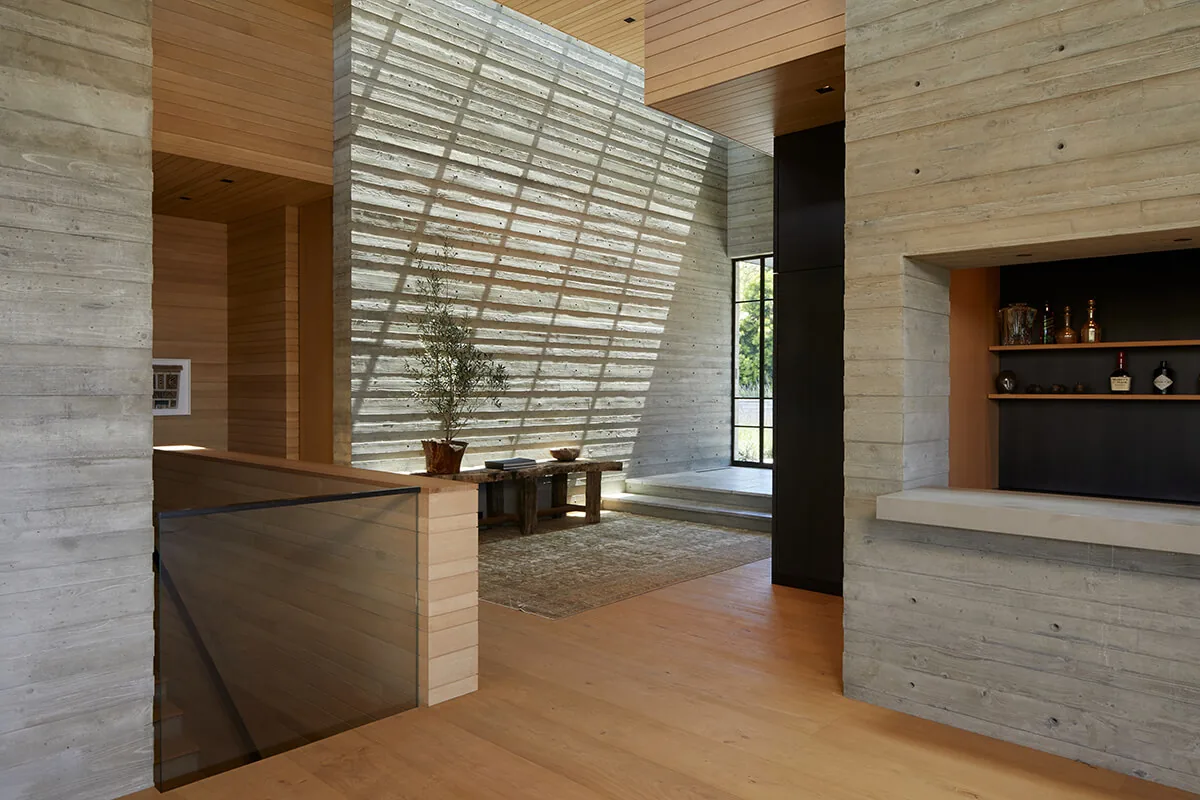 Living area of the Sapire Residence features exposed board-formed concrete walls and wood paneling with a rustic bench and potted olive tree.