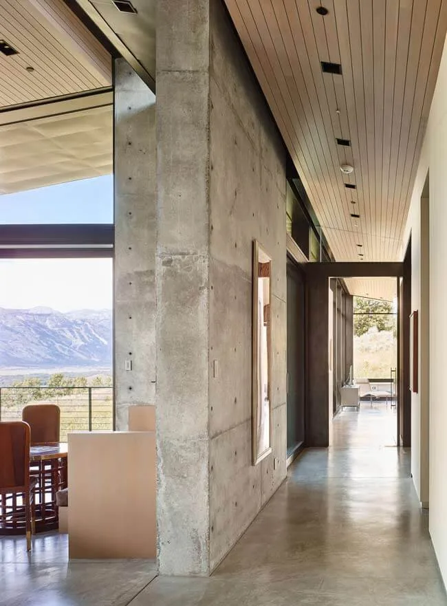 Interior hallway of Wyoming Residence features polished concrete floor, wood plank ceiling, and a concrete accent wall with art near large windows