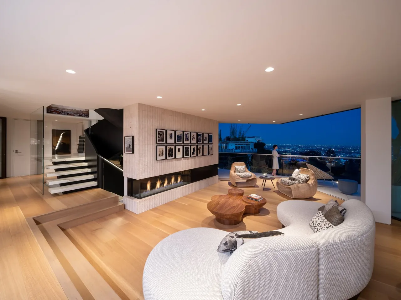 Living room with curved sofa, floating open-riser staircase, linear fireplace, and floor-to-ceiling glazing showing city views at dusk.
