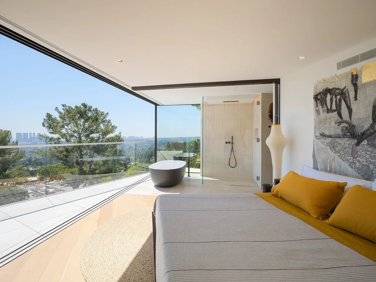 Bedroom with floor-to-ceiling glass walls overlooking a forested landscape, connected to a bathroom with a freestanding tub and a glass shower enclosure.