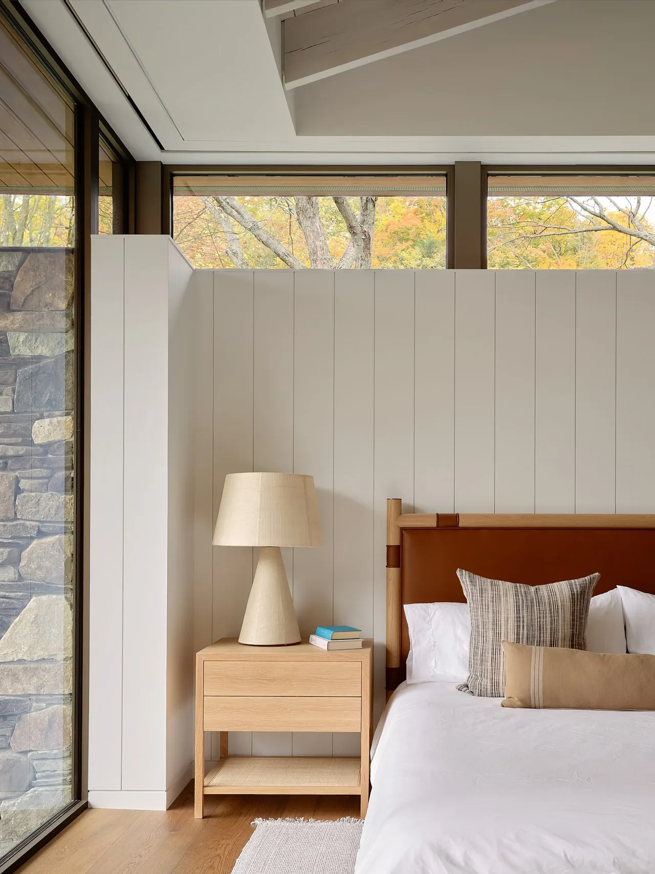 Bedroom corner in Muskoka Cottage with a leather headboard bed and natural wood nightstand topped with a lamp and books.