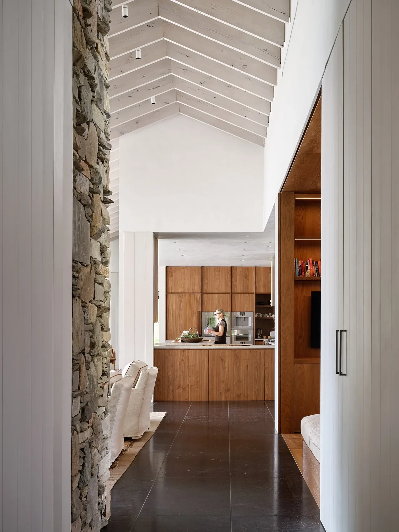Vaulted hallway leads to a kitchen featuring wood cabinetry and a large island with a person preparing food.