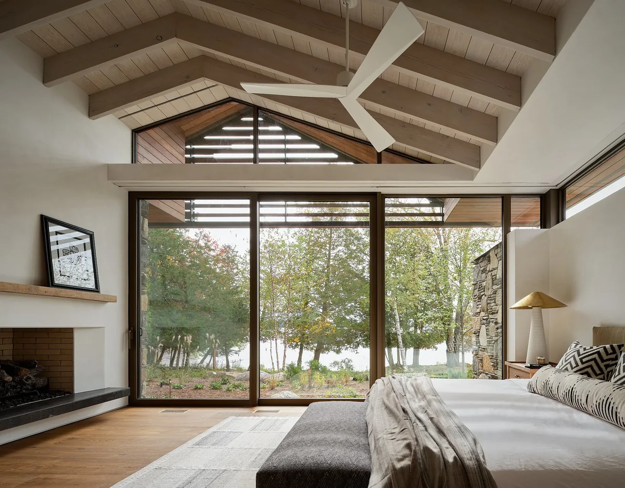 Bedroom features a vaulted ceiling with exposed beams, a fireplace, and expansive glass doors to a lakeside view.