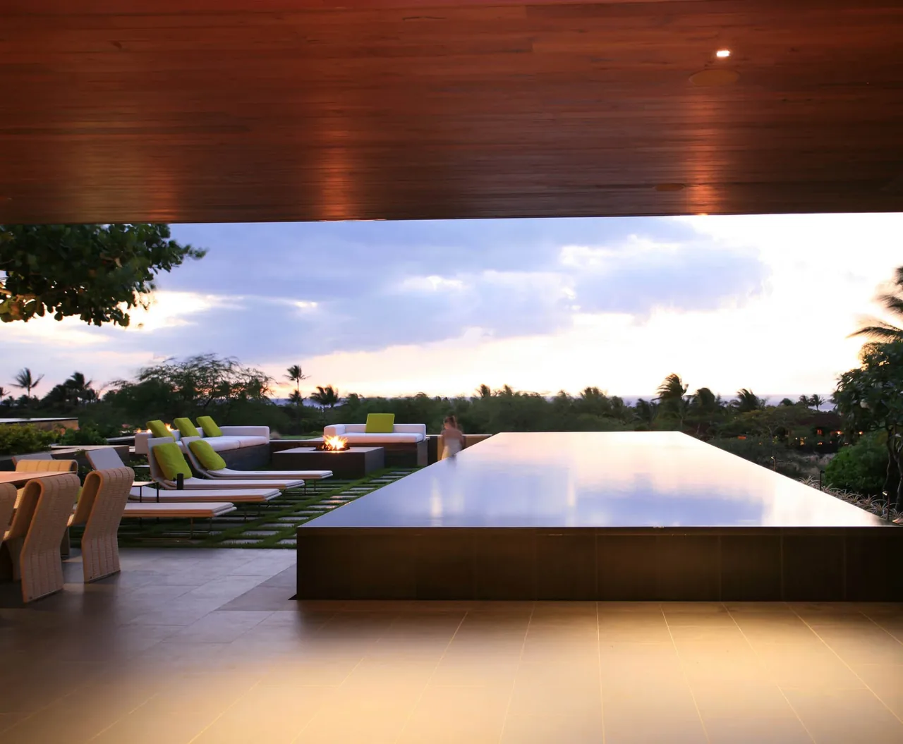 A covered patio features recessed downlights, a reflecting pool, outdoor seating with cushions, and landscaping with palm trees at sunset.