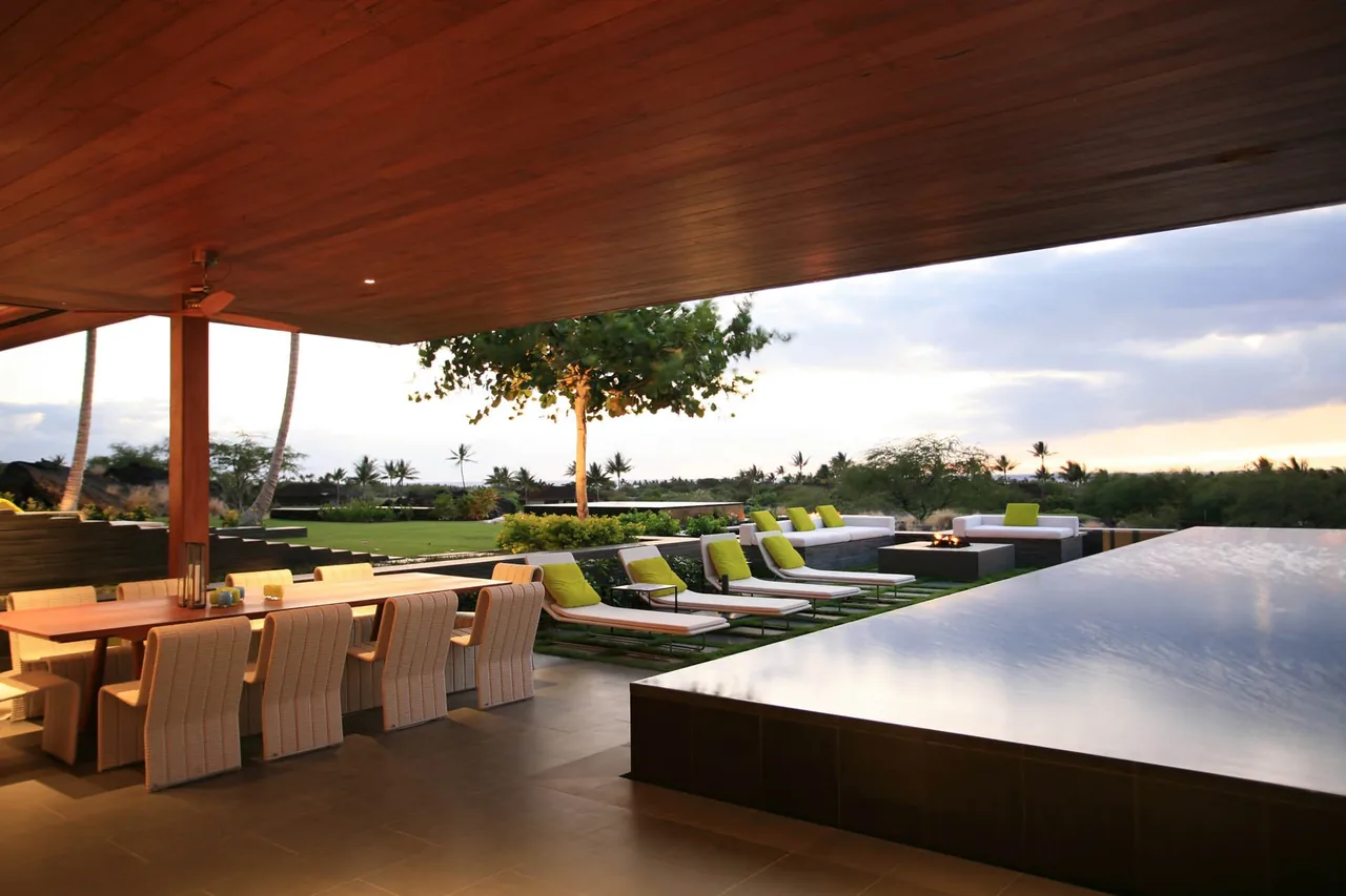 Covered patio with dining area, lounge chairs, and fire pit next to an infinity-edge pool overlooking landscaped greenery.