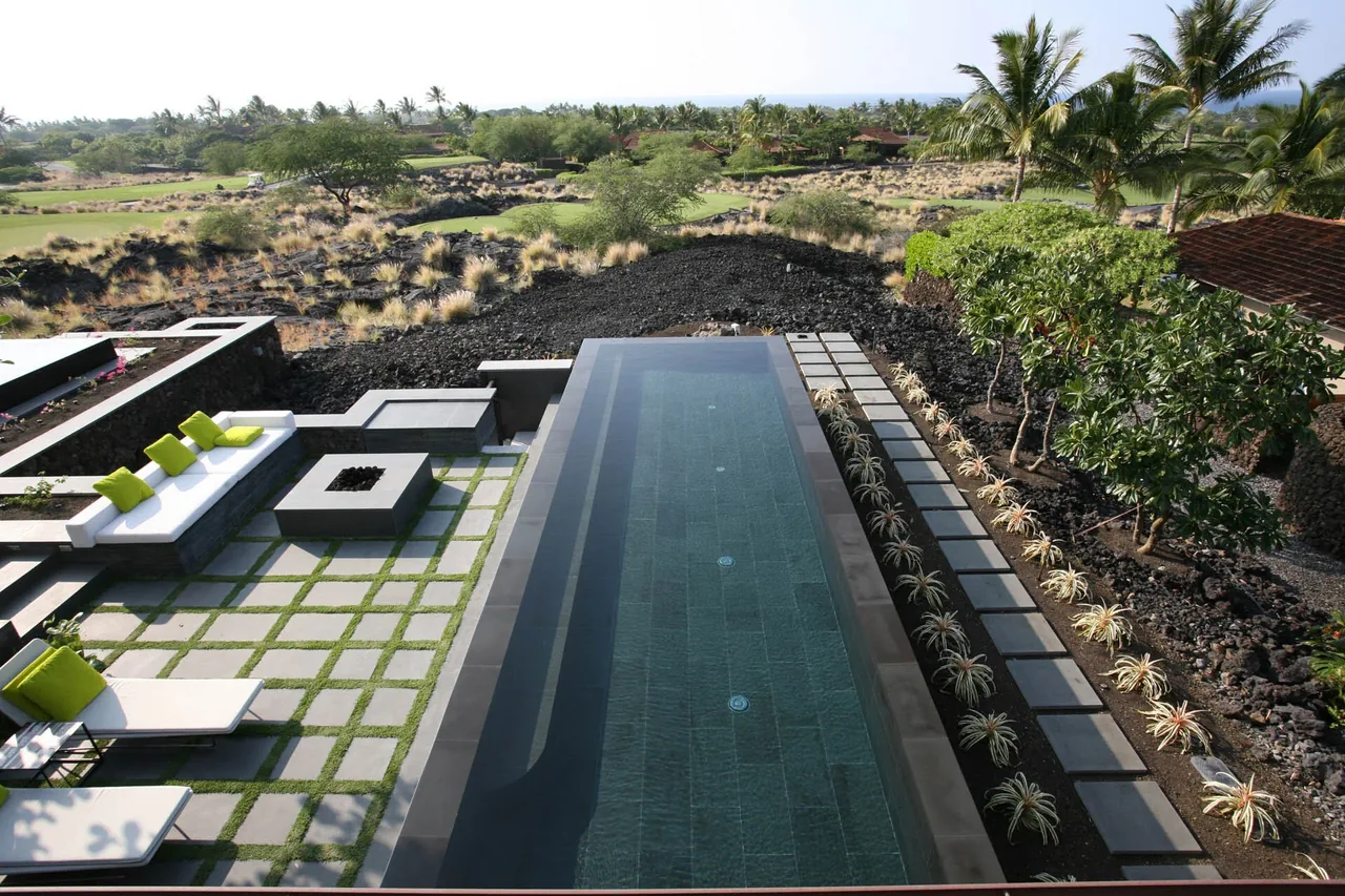 Outdoor patio area with a long rectangular pool, concrete tile flooring with grass in between, built-in seating with green cushions, and landscaping with plants and palm trees.