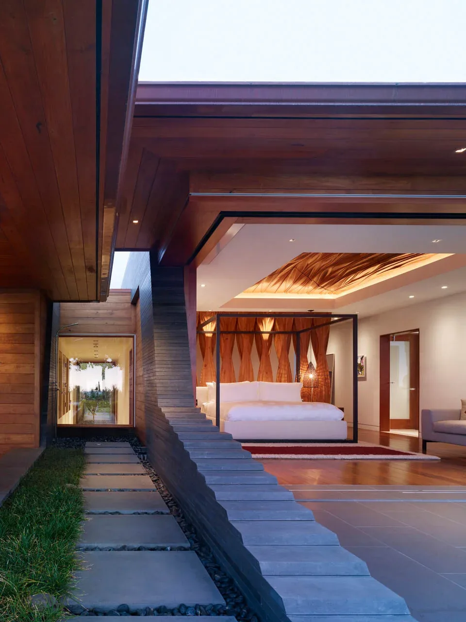 Modern bedroom opens to an outdoor walkway with stone pavers beside a grass strip, featuring wood ceiling and concrete steps.
