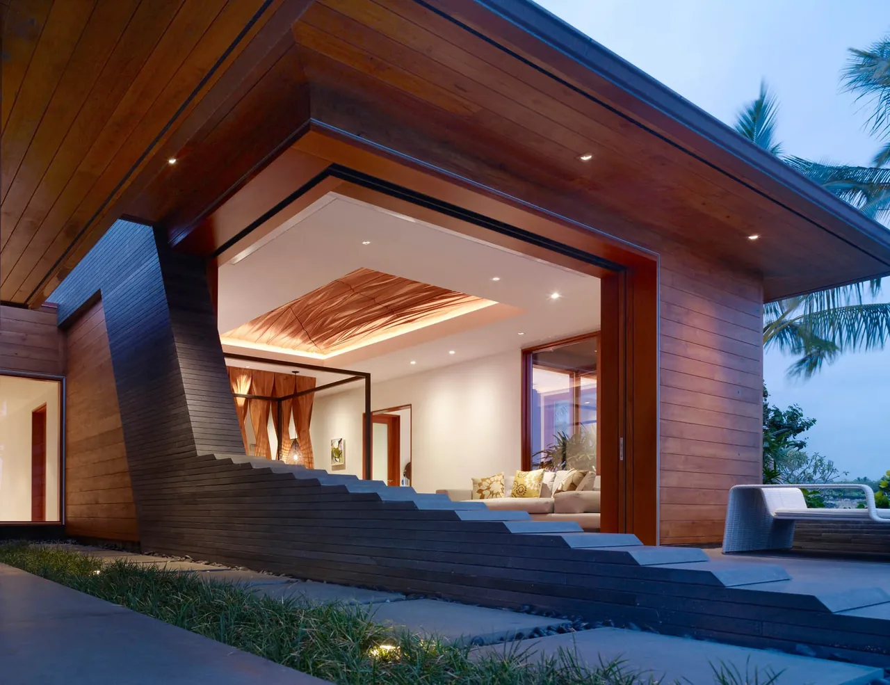 Modern residence exterior features stepped concrete wall, wood-clad covered patio, and built-in lighting at dusk.