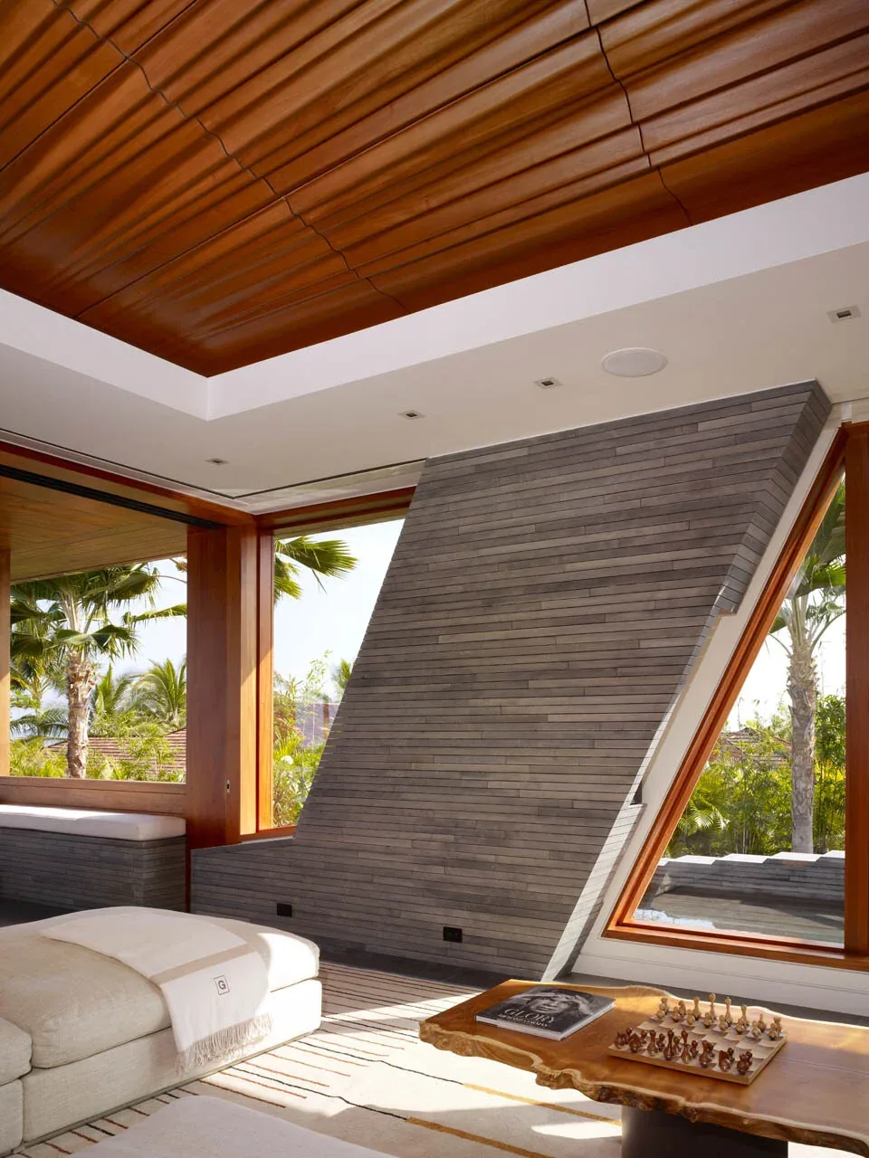 Living room interior with gray tiled angled wall, wood-framed large windows, and a wood paneled ceiling.