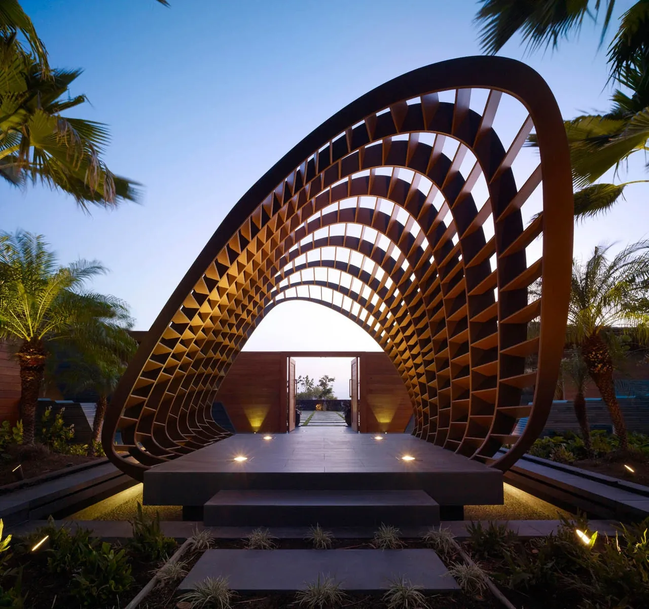Curved wooden lattice arch forms an architectural focal point over an elevated concrete platform in an outdoor garden setting.