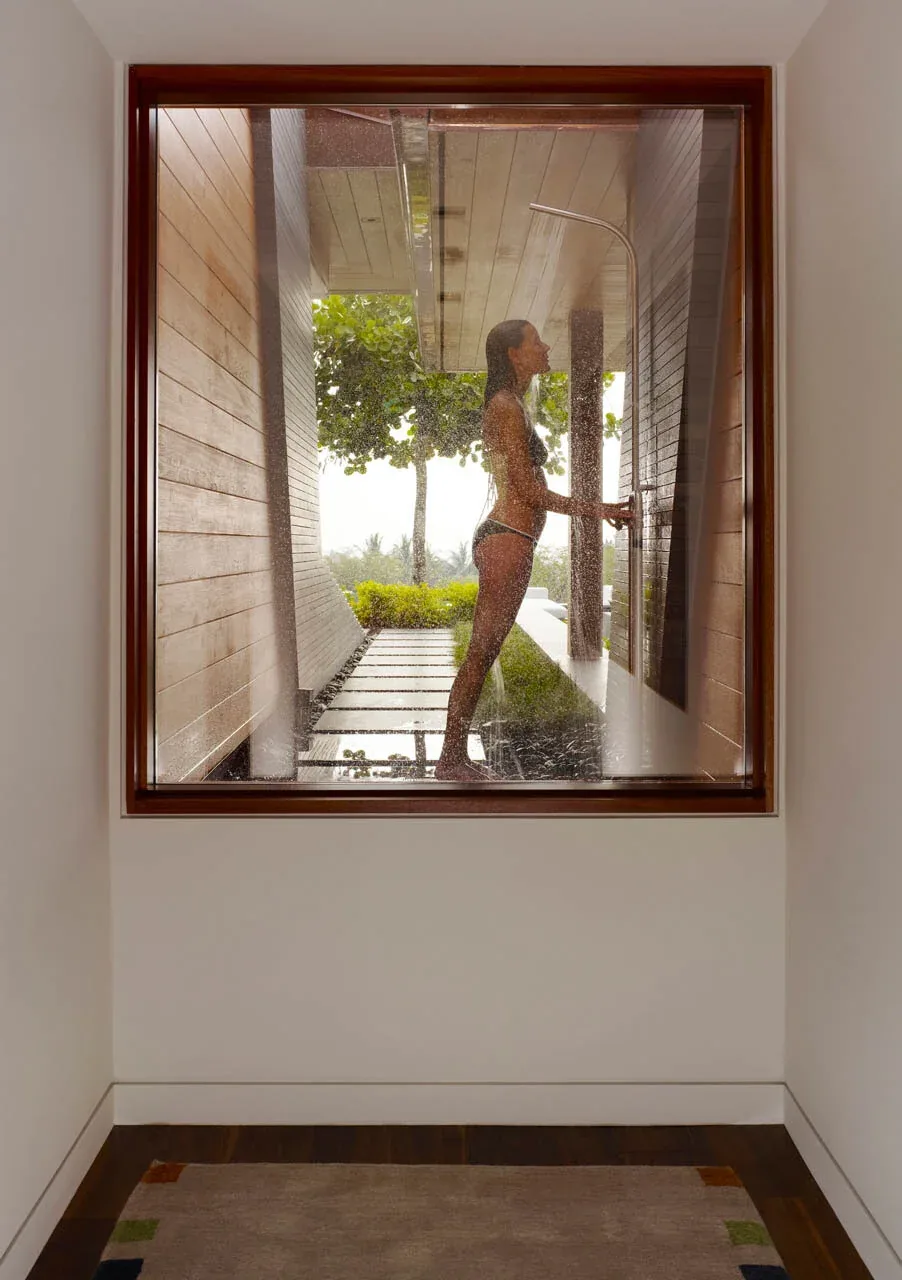 Exterior corridor with outdoor shower viewed through floor-to-ceiling glazing and wood framing.