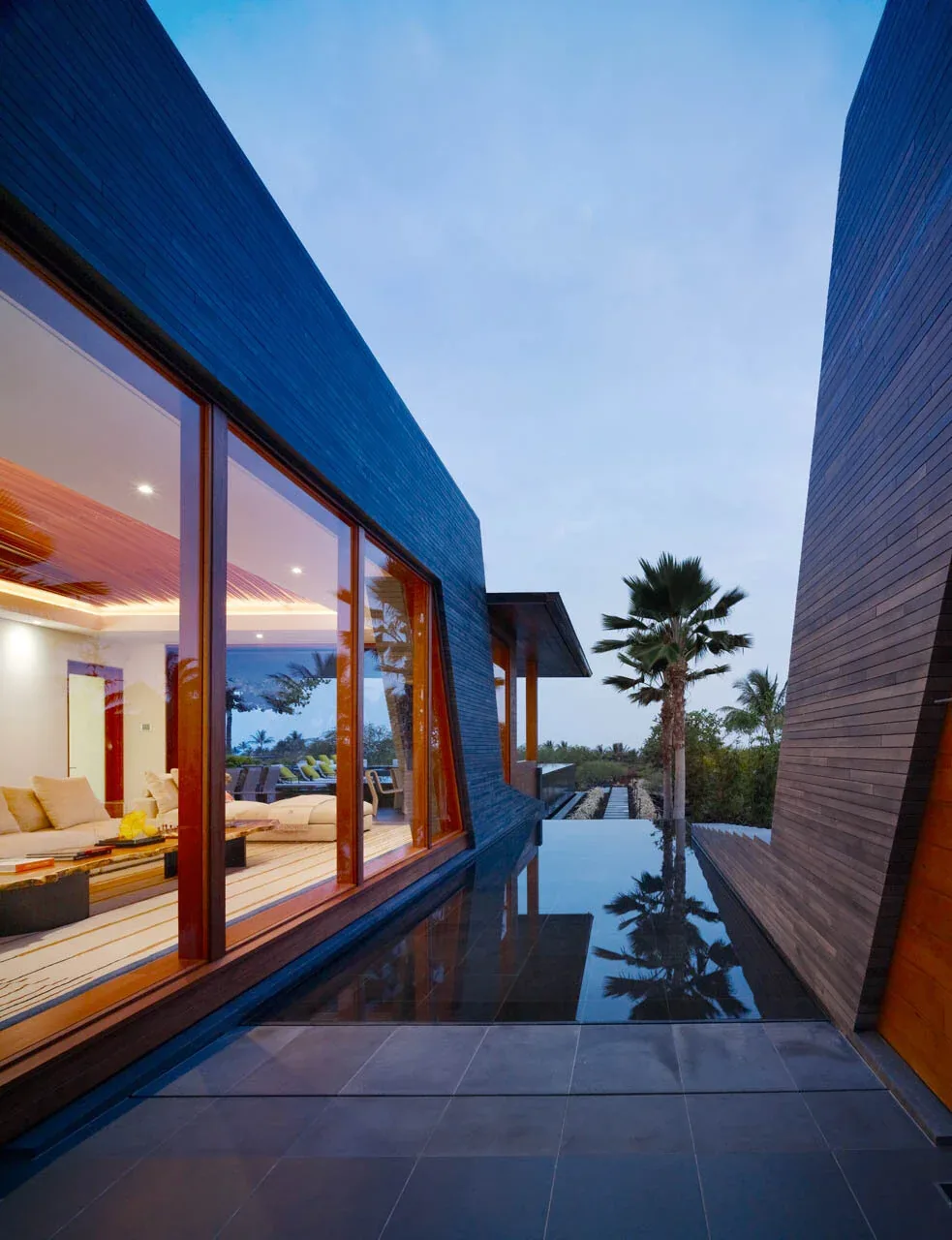 Exterior side view of a residence featuring reflective pool water bordered by dark wood siding walls and large windows revealing a sofa inside.