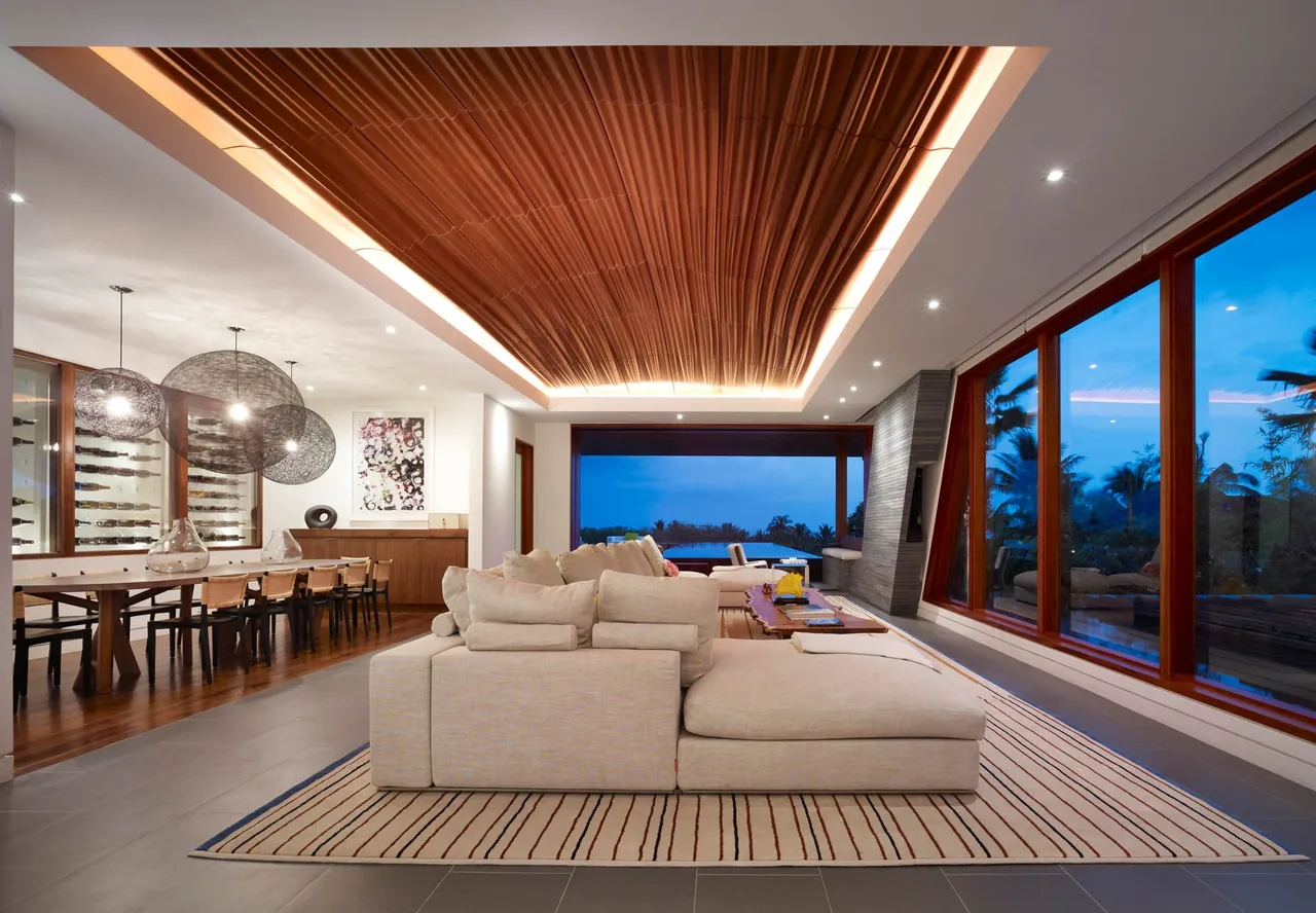 Open living and dining room with wood ceiling feature and large glass windows looking out to vegetation during dusk.