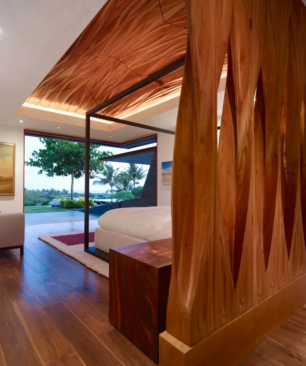 Bedroom with wooden floor, sculptural wooden wall divider, canopy bed, and large sliding glass door opening to outdoor landscape.