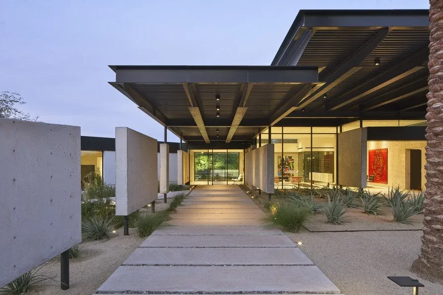 Walkway with concrete panels and a roof structure leading to a glass entrance surrounded by desert landscaping at dusk.