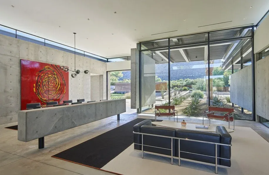 The Camelback Residence features a contemporary living room with concrete half-walls and floor-to-ceiling glazing overlooking desert vegetation.