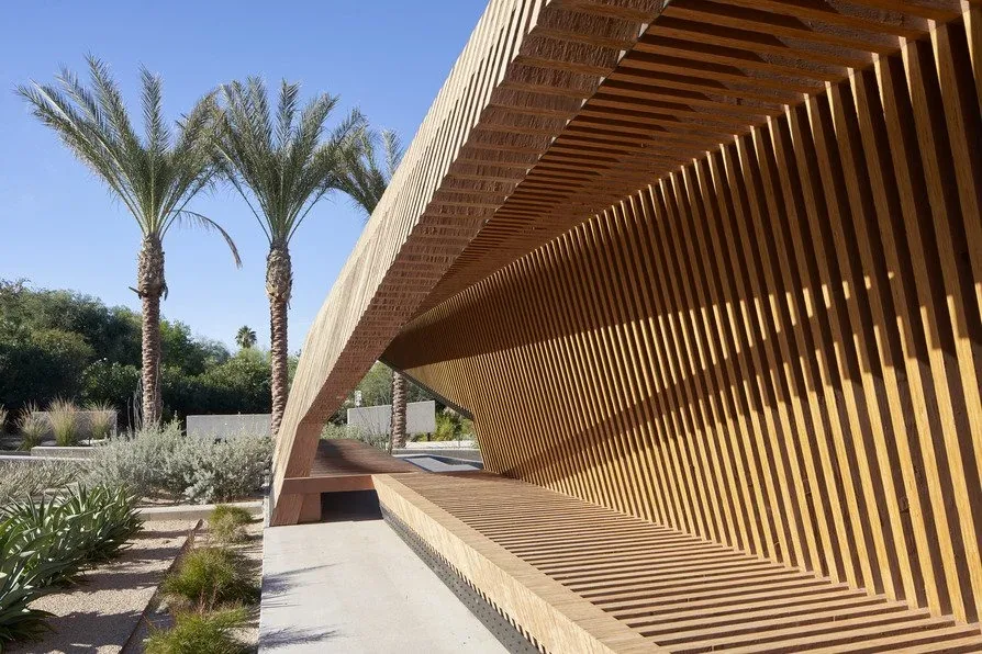 Outdoor seating framed by wooden slats casting patterned shadows in a desert landscape with palm trees.