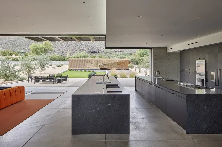 Open kitchen in a modern desert home features black stone islands and floor-to-ceiling windows opening to an outdoor patio and garden.