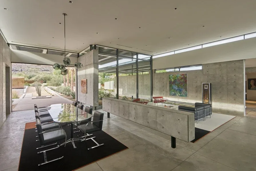 Camelback Residence features concrete interior walls, floor-to-ceiling glazing, and a glass dining table with black chairs.