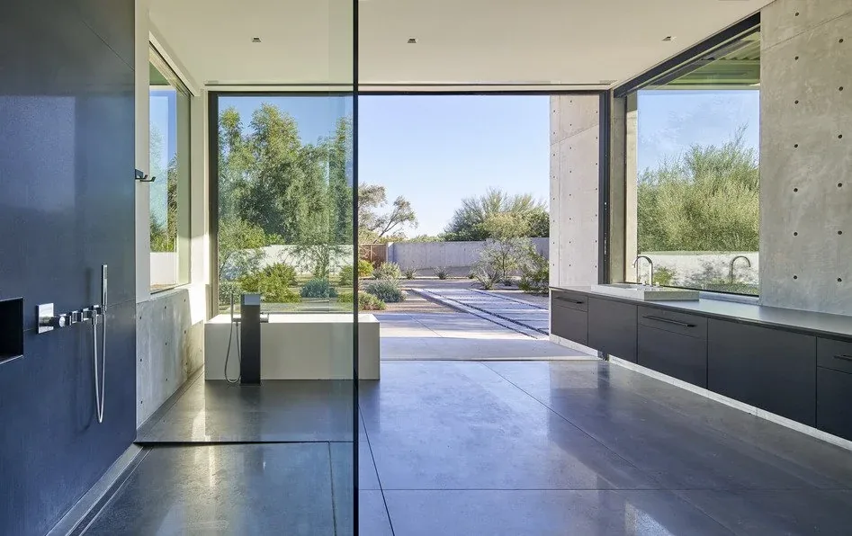 Bathroom with concrete floors and walls, floor-to-ceiling glazing, and black cabinetry overlooking desert vegetation.