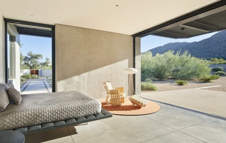 Open bedroom with floor-to-ceiling glazing and concrete walls overlooks desert vegetation and mountains.