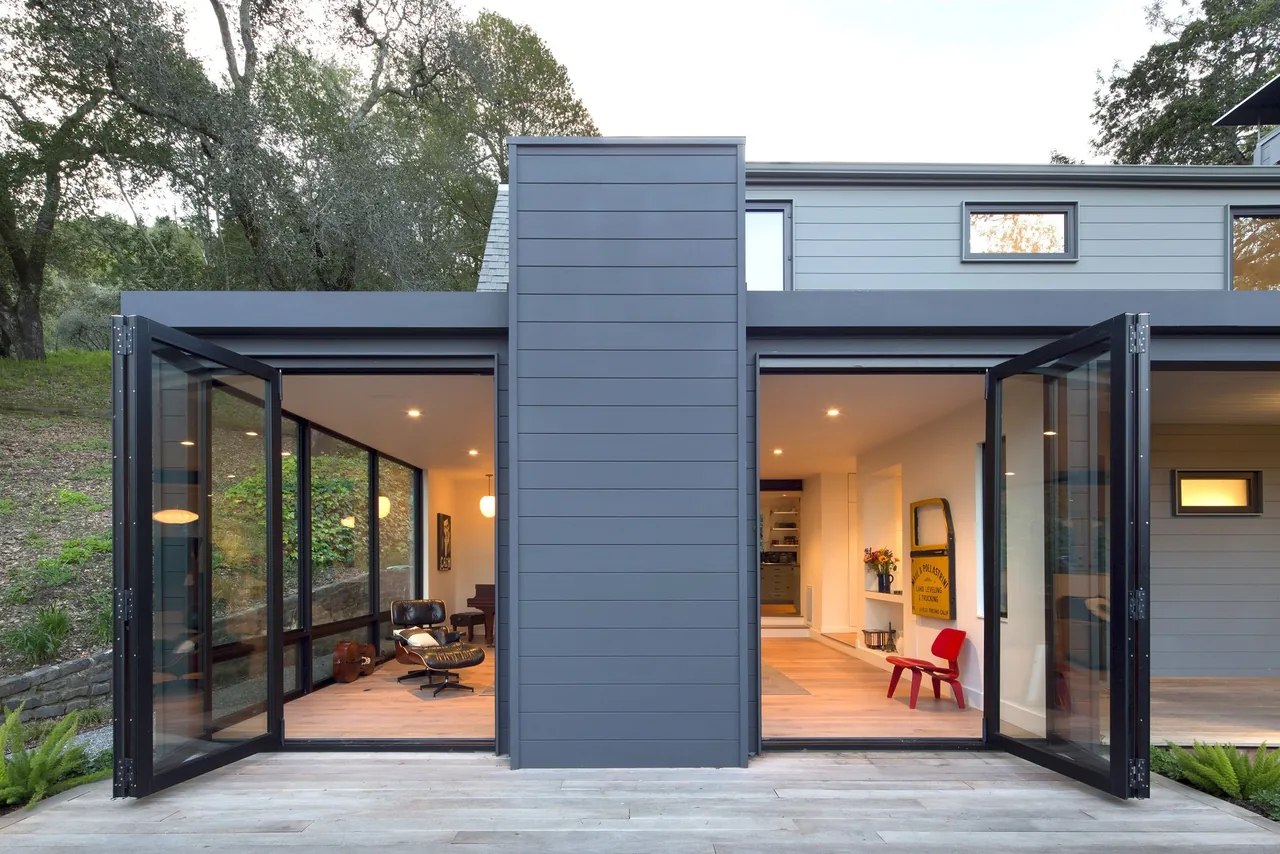 Modern barn-style home features black-framed folding glass doors opening to a wood deck and hillside view.