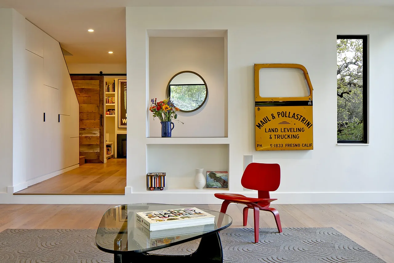 Modern living room featuring light oak flooring, built-in wall niches, and an artistic yellow truck door mounted on a white paneled wall.
