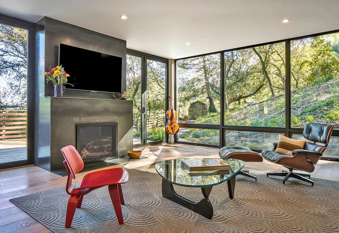 Living room with corner windows and a metal fireplace wall featuring a mounted TV, overlooking a hillside landscape.