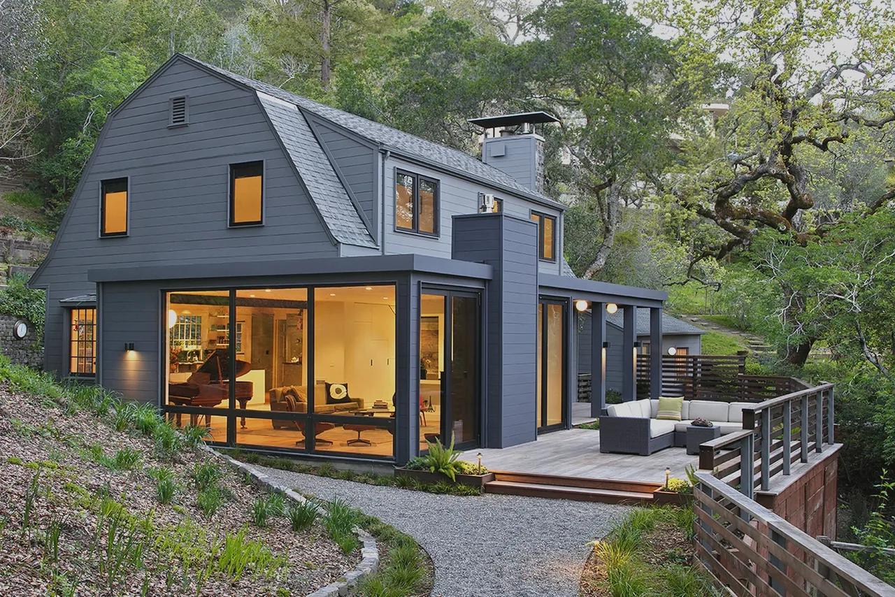 Modern barn-style house with gray siding, large floor-to-ceiling windows, and a wood deck on a hillside.