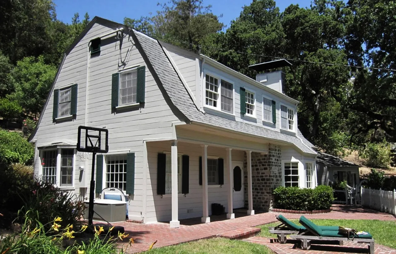 Traditional two-story house with gambrel roof, white siding, green shutters, and brick patio with outdoor furniture.