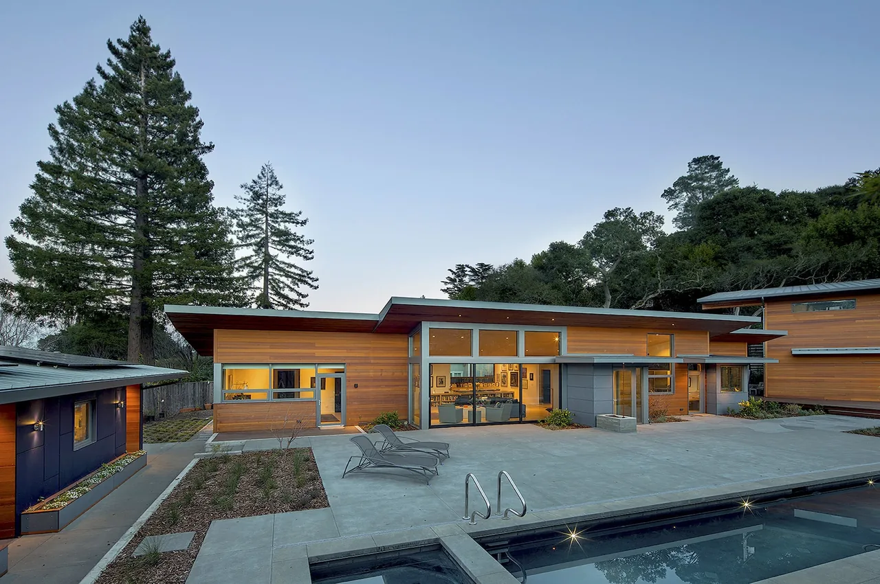 The Net Zero Energy House features wood siding and expansive windows opening to a concrete poolside patio.
