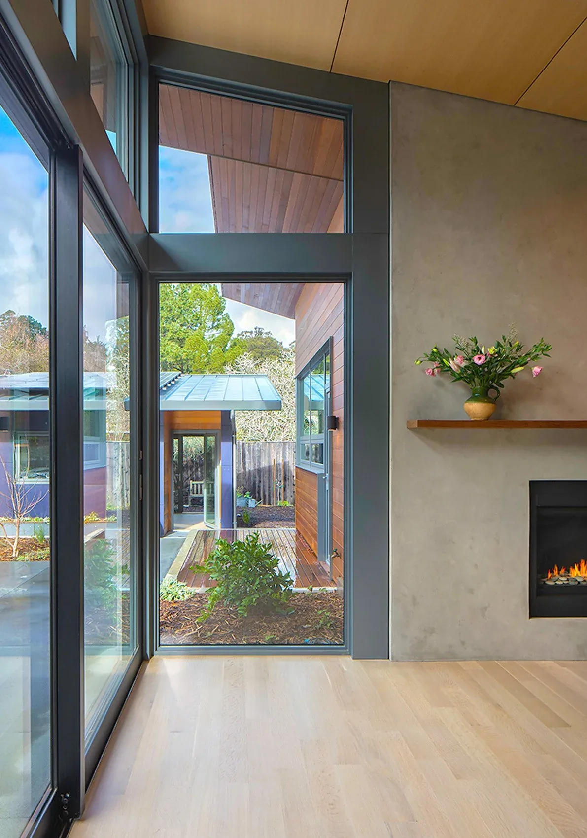 Modern living room with floor-to-ceiling glazing, wood ceiling panels, and a linear fireplace beside a garden view.