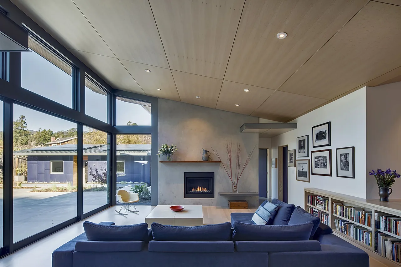 Living room features a concrete fireplace, wood-paneled ceiling, and floor-to-ceiling glazing with black window frames.
