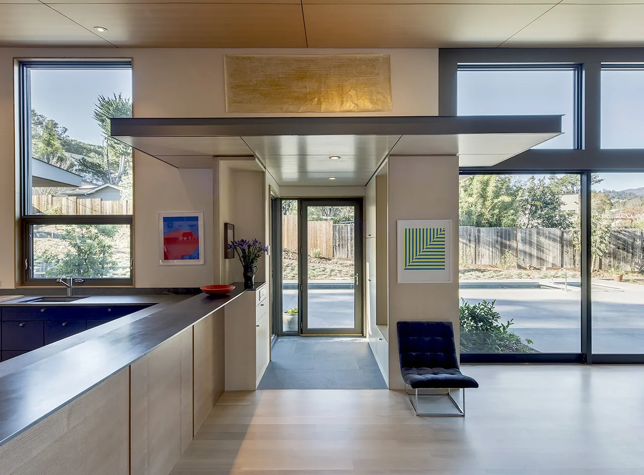 A modern kitchen space featuring large metal-framed windows and a glass door with wood ceiling panels.