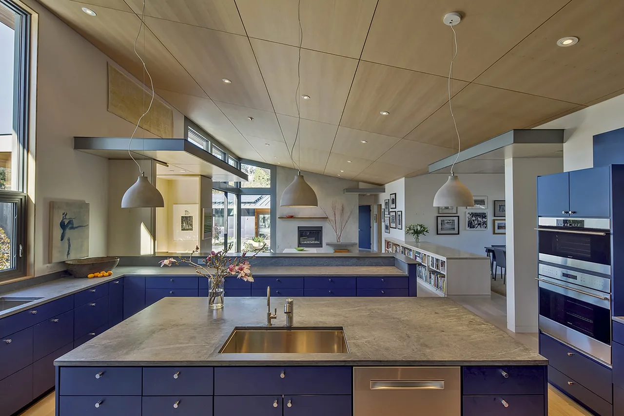 Kitchen with blue cabinets, concrete counters, wood ceiling panels, and pendant lighting in the Net Zero Energy House project.