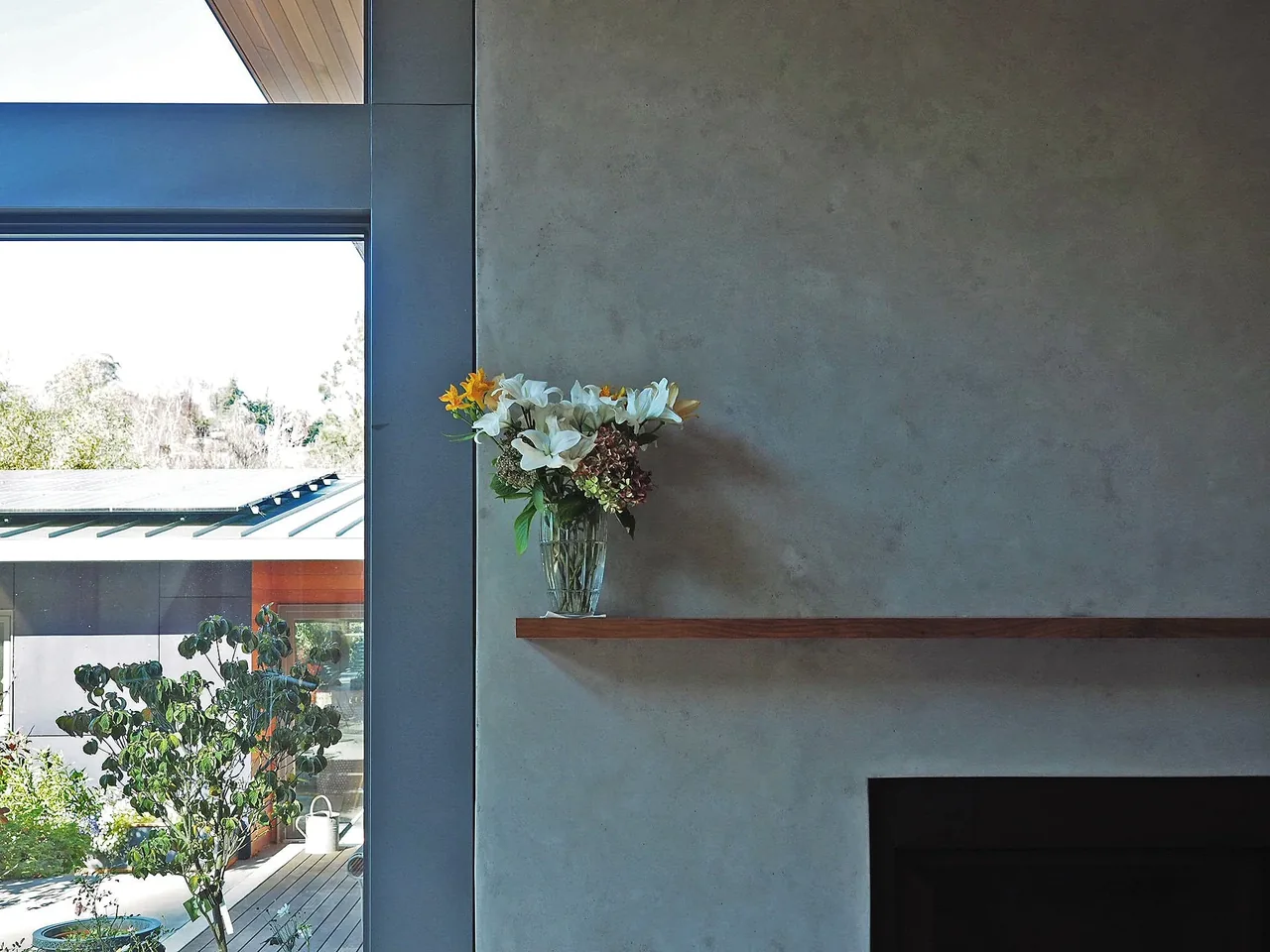 A minimalist fireplace with a wooden mantel holding a vase of flowers and a large window showing a landscaped exterior.