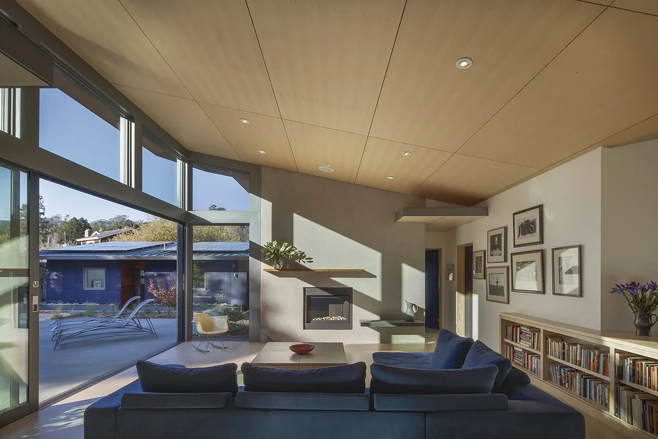 A modern living room in the Net Zero Energy House with wood ceiling panels, concrete fireplace wall, and poolside views.