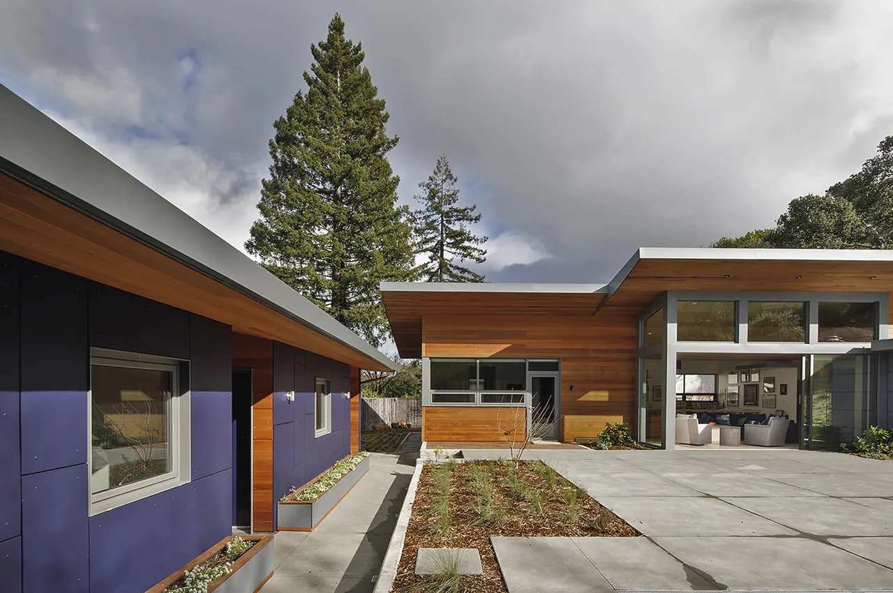 Modern house exterior showing wood siding, large metal windows, and concrete patio with landscaped beds.
