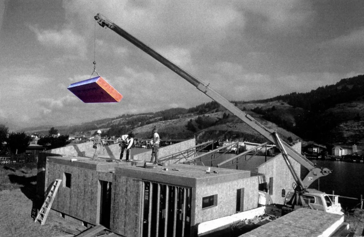 Workers install a section of the Sail Loft House wall with a crane, overlooking waterfront and hillside views.