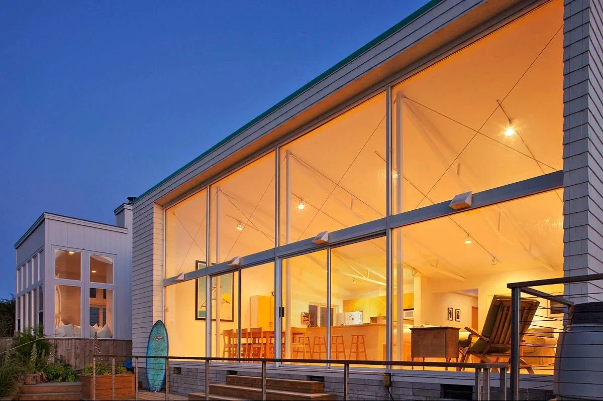 Sail Loft House features expansive floor-to-ceiling glazing that reveals a warm, open living and kitchen space at dusk.