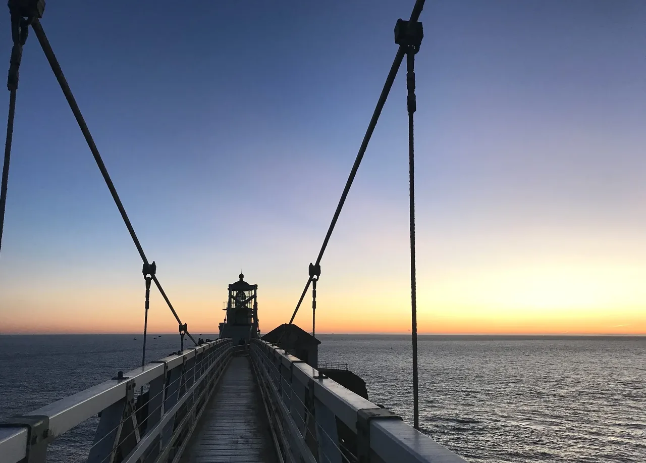 Pier walkway with wire railing leading to lighthouse at dusk with waterfront view.