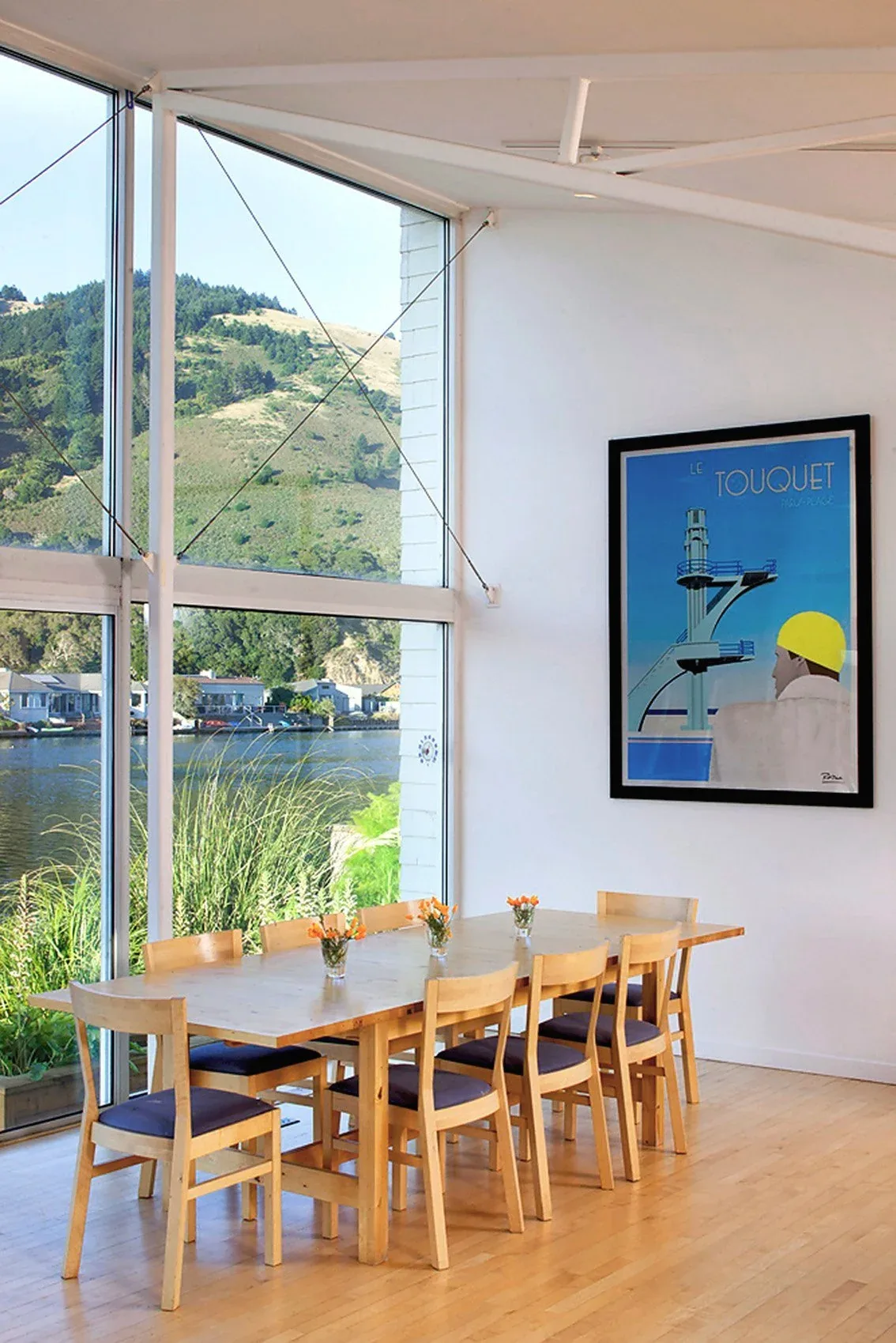 Dining space featuring a wooden table with chairs adjacent to floor-to-ceiling glazing with a view of water and hills.
