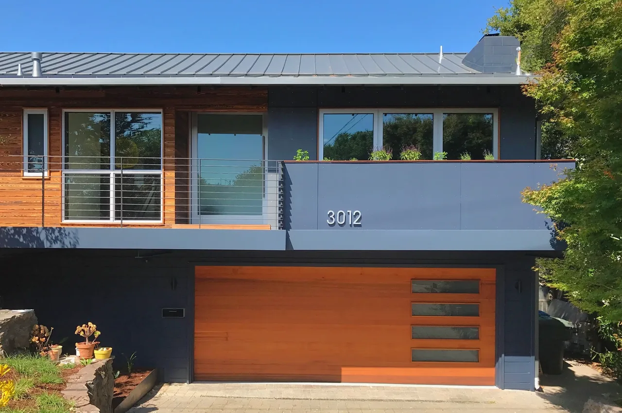 Modern house exterior with a metal roof, wood garage door, and balcony featuring a horizontal railing.