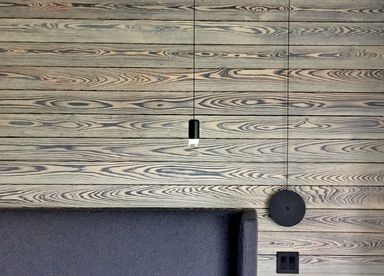 Interior wall features horizontally arranged wooden planks with distinct grain and a black and white pendant light over gray seating.
