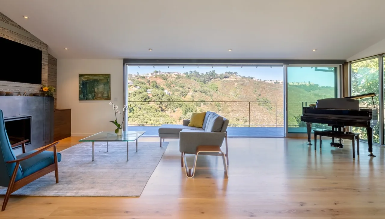 Living room features light wood floor, expansive glass window wall with views, and minimal modern furniture.